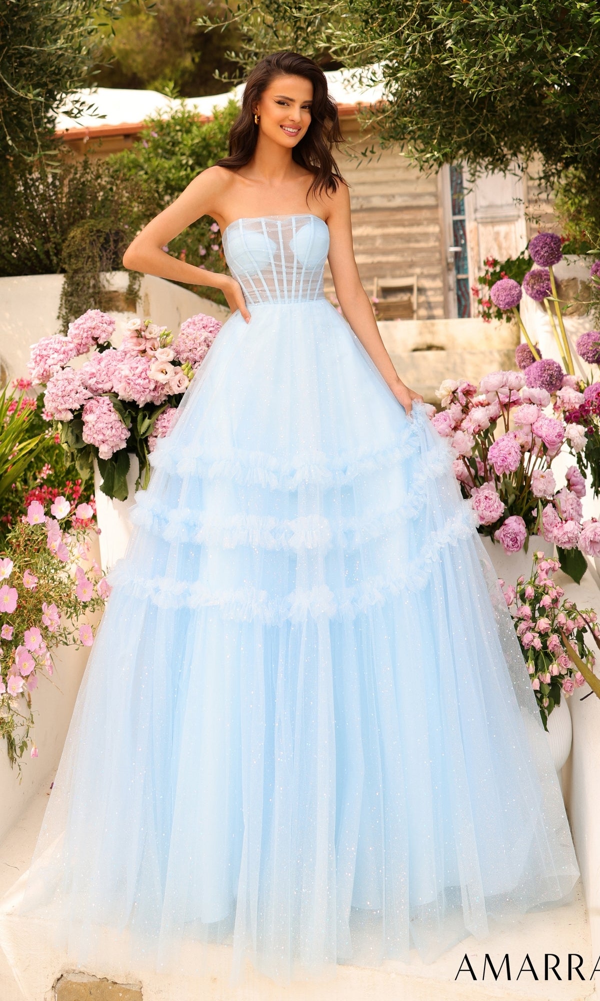 A woman stands outdoors among pink flowers, wearing the Amarra Strapless Sheer-Bodice Prom Ball Gown 88794 in light blue with a fitted bodice and layered glitter-tulle overlay skirt.